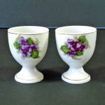 A pair of white porcelain egg cups decorated with purple violets on both sides. They're 2-1/4" high by 1-3/4 inches across the rims. There are no marks, my guess would be they were made in Mid Ce...